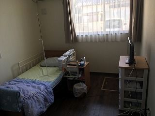 寝具付きのベッドや収納ケースが置かれた部屋にはレースカーテンとカーテンが付いた大きな窓がある。