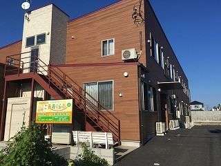 木目調の２階建ての建物は玄関には屋根があり、屋外階段のそばには白いベンチが置かれている