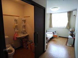 大きなスライド式のドアが付いたトイレが居室内にあり、窓際には寝具付きのベッドが置かれている。