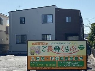 グレー系の2階建ての建物は広い敷地内にあり、駐車スペースもある。手前にはホームと併設事業所の看板もある。