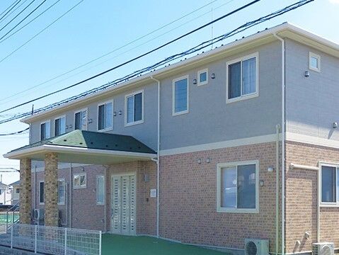 2階建ての建物はブラウン系で建物の周りには白いフェンスが立ち、玄関部分には屋根が付いている。