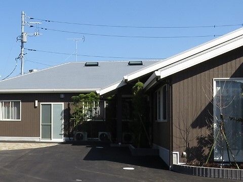 ブラウンの平屋建ての建物にはグレーの三角屋根が付き、敷地内にはアスファルトが敷かれ、緑も植えられている。