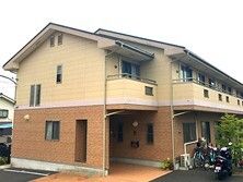 ２階建ての建物は敷地内に草木が植えられ、ベージュとブラウンの外壁。2階部分にはベランダがある。