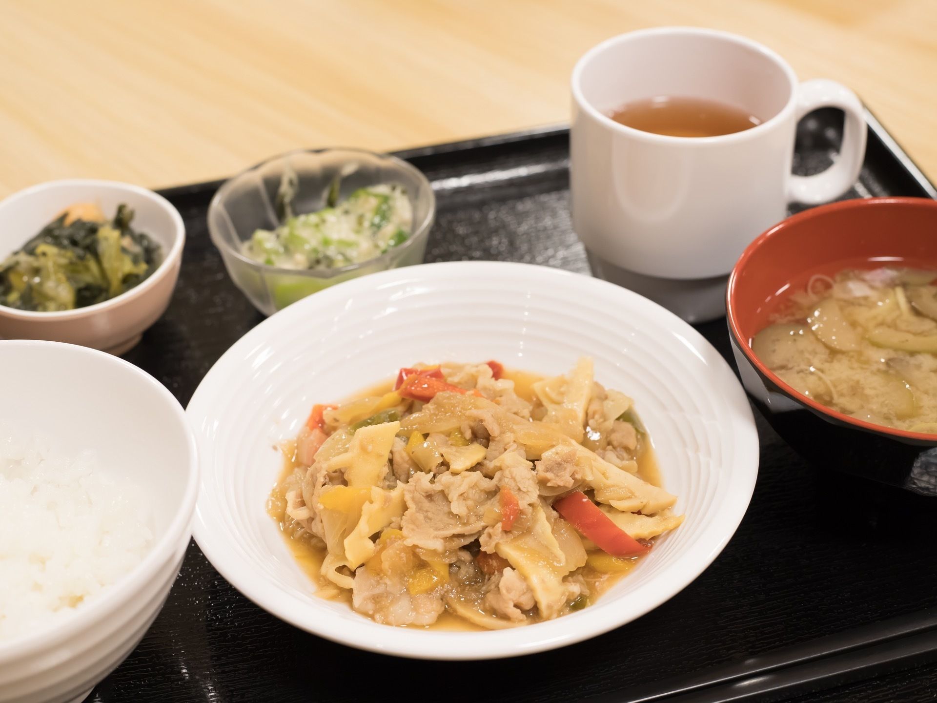黒いトレーの上には、5種類の器に盛られた食事が並べられている。右側には、白色のお茶の入ったコップが置かれている。