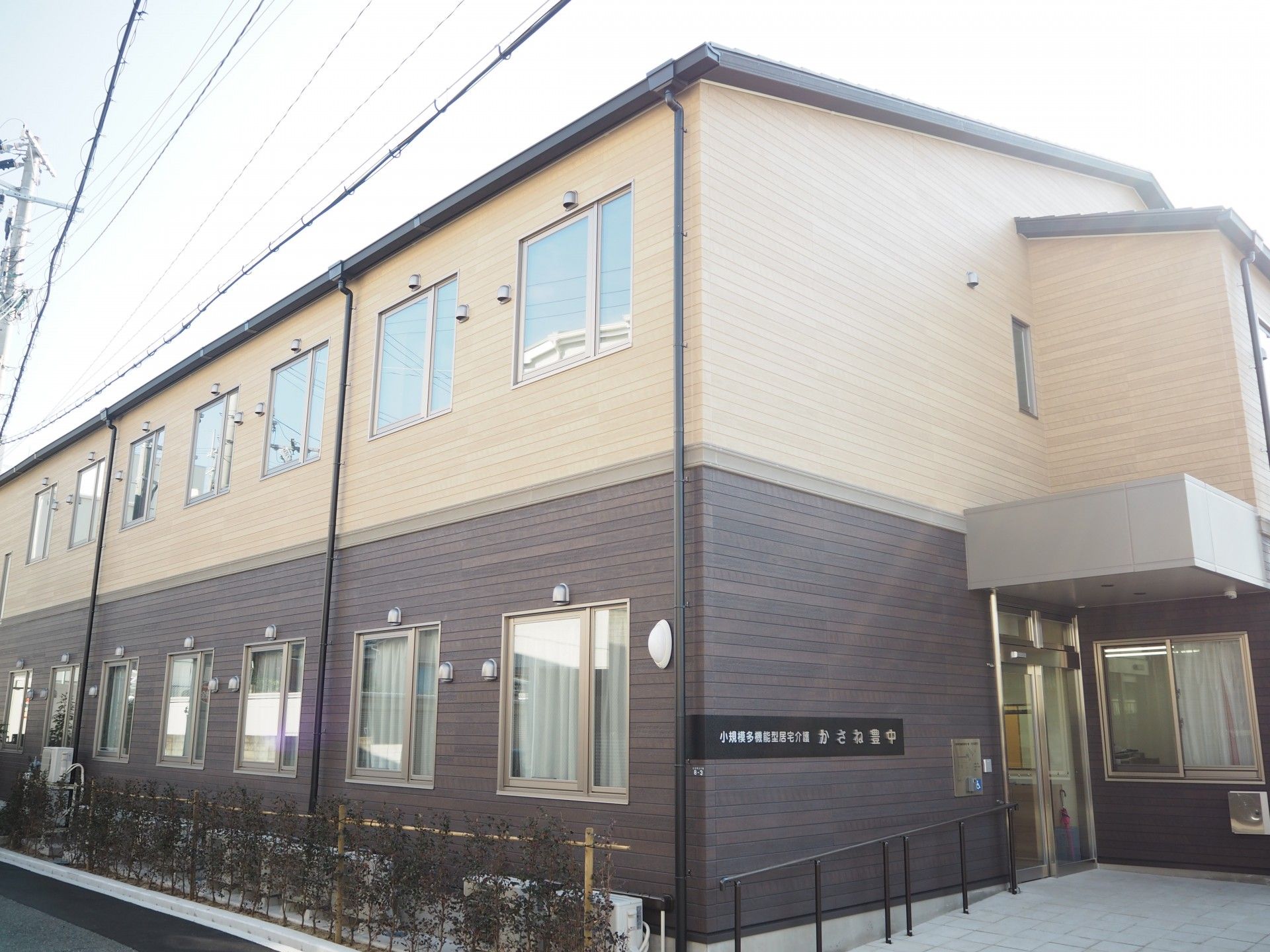 2階建ての建物。1階部分はブラウン、2階部分はベージュで、生垣もある。
