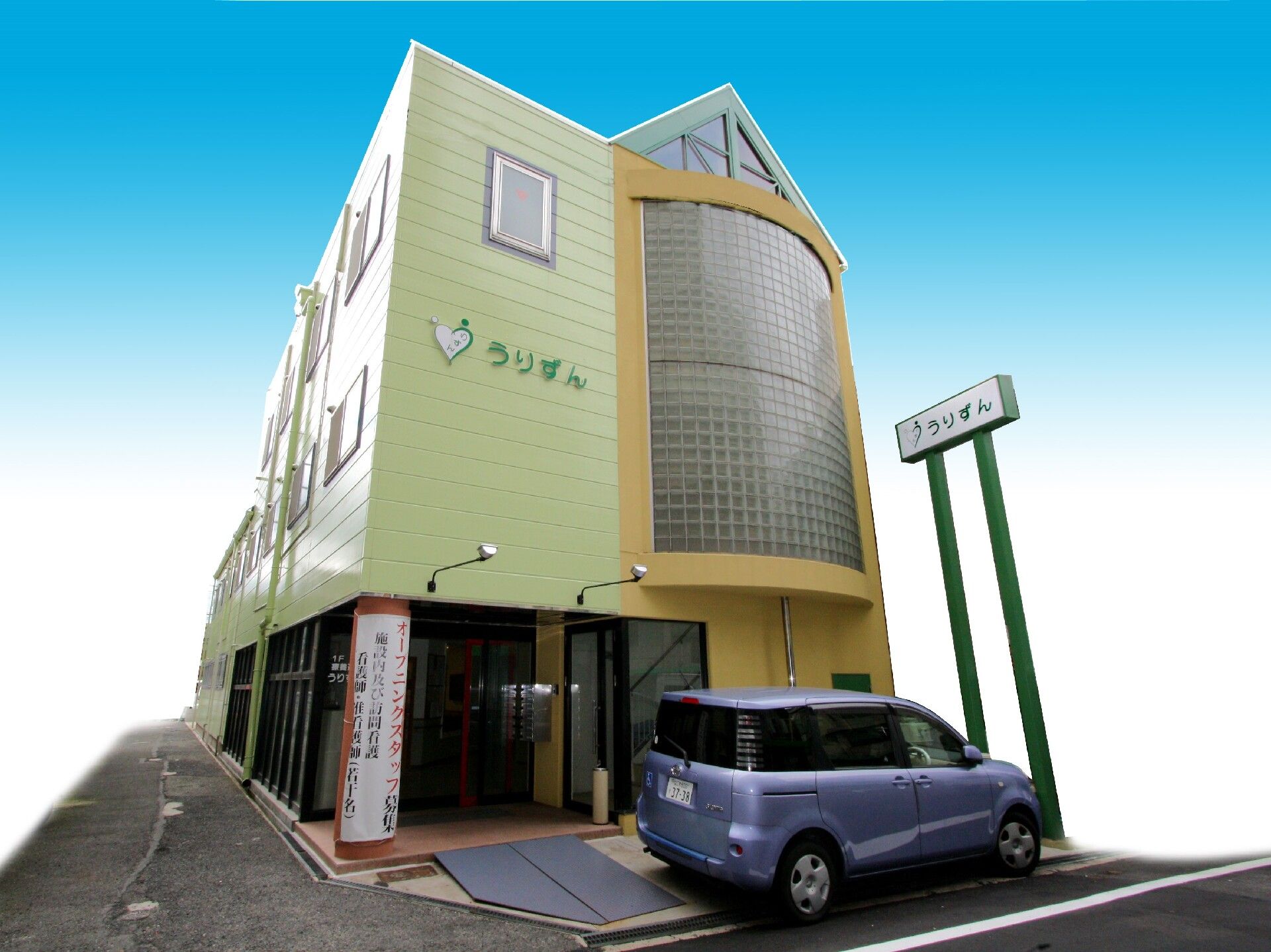ライトグリーンとイエローの4階建ての建物の玄関の前には1台の車が止められていて、外壁にはホーム名が書かれている。