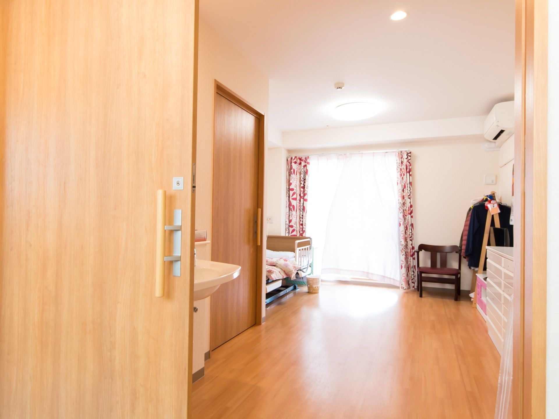 木製の引き戸の向こうにフローリングの部屋がある。窓の近くにベッドがあり、反対側には白いチェストや衣装掛けが置かれている。