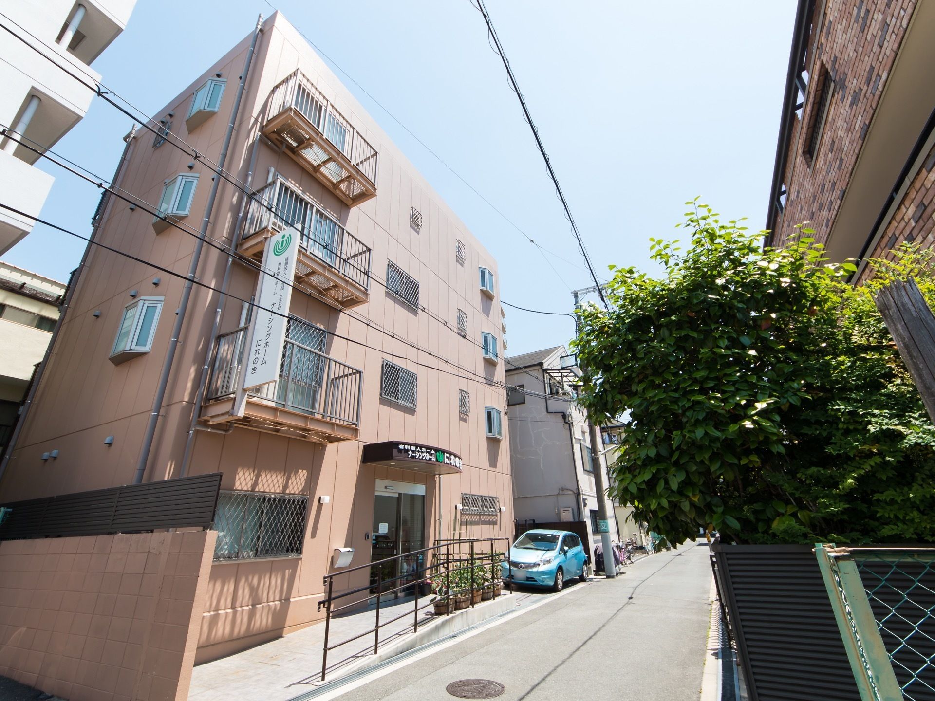 路地に4階建ての建物が建っている。周囲は同じような高さの集合住宅が並んでいる。車道から玄関までは手すり付きのスロープになっている。