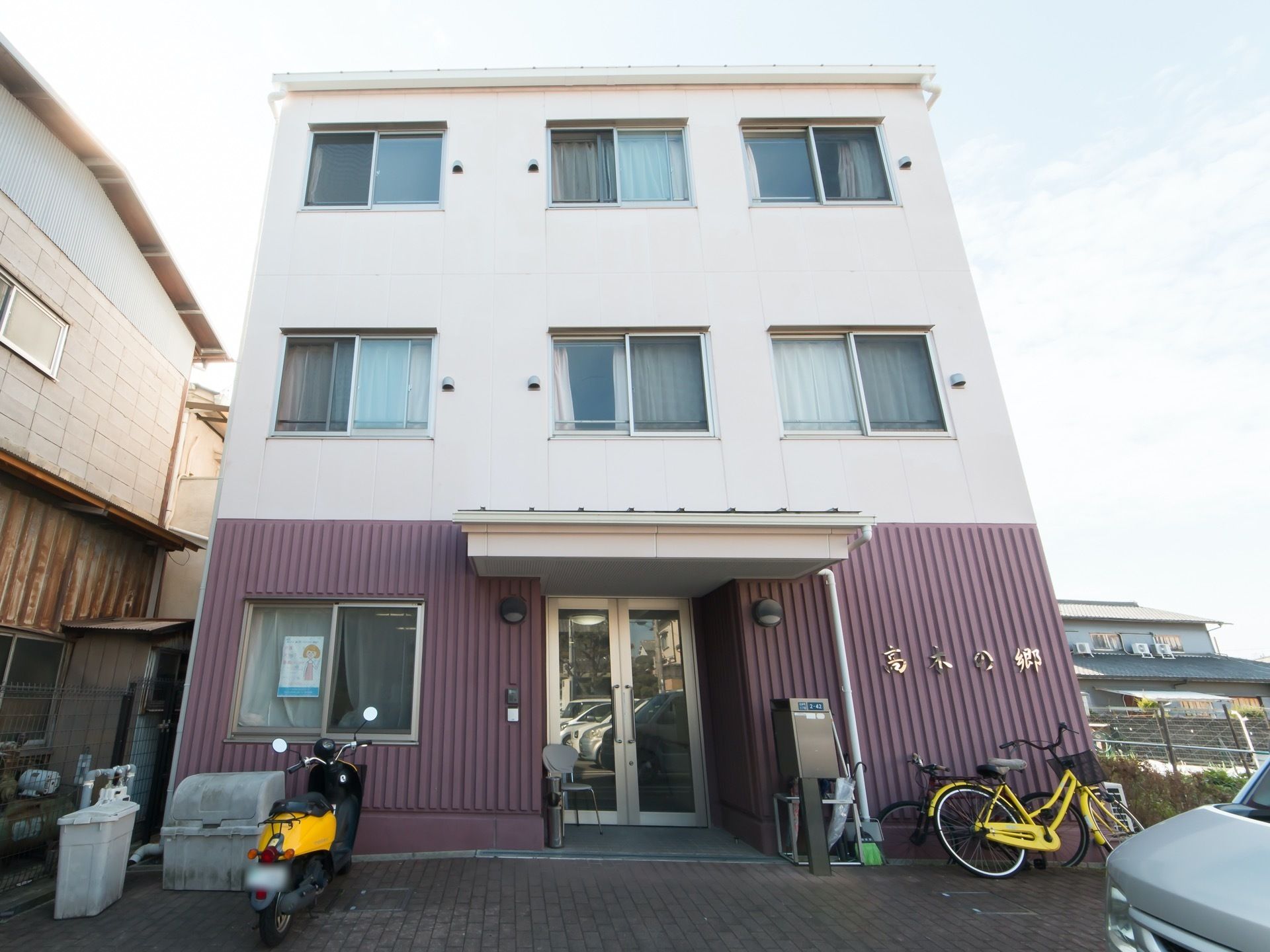 ツートーンの外壁が特徴的な３階建ての建物である。目の前に駐車場があり、自転車やオートバイが置かれている。玄関には広い屋根が備わっている。