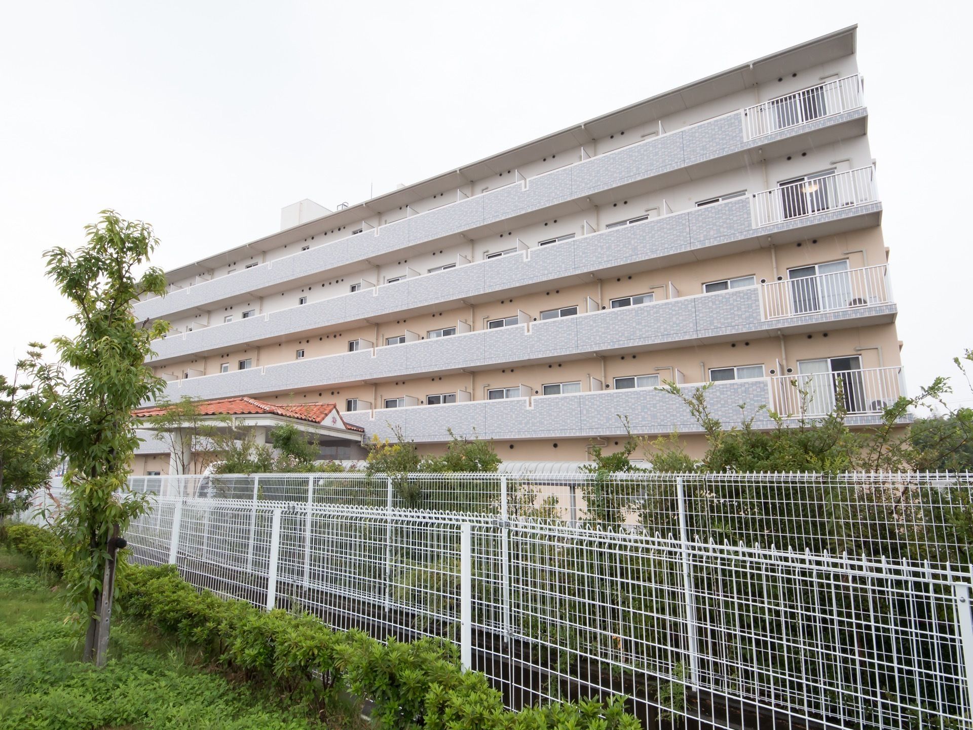 ５階建て施設は、薄ピンク色の外壁で統一されている。敷地の周辺には、白いフェンスが取りつけられている。
