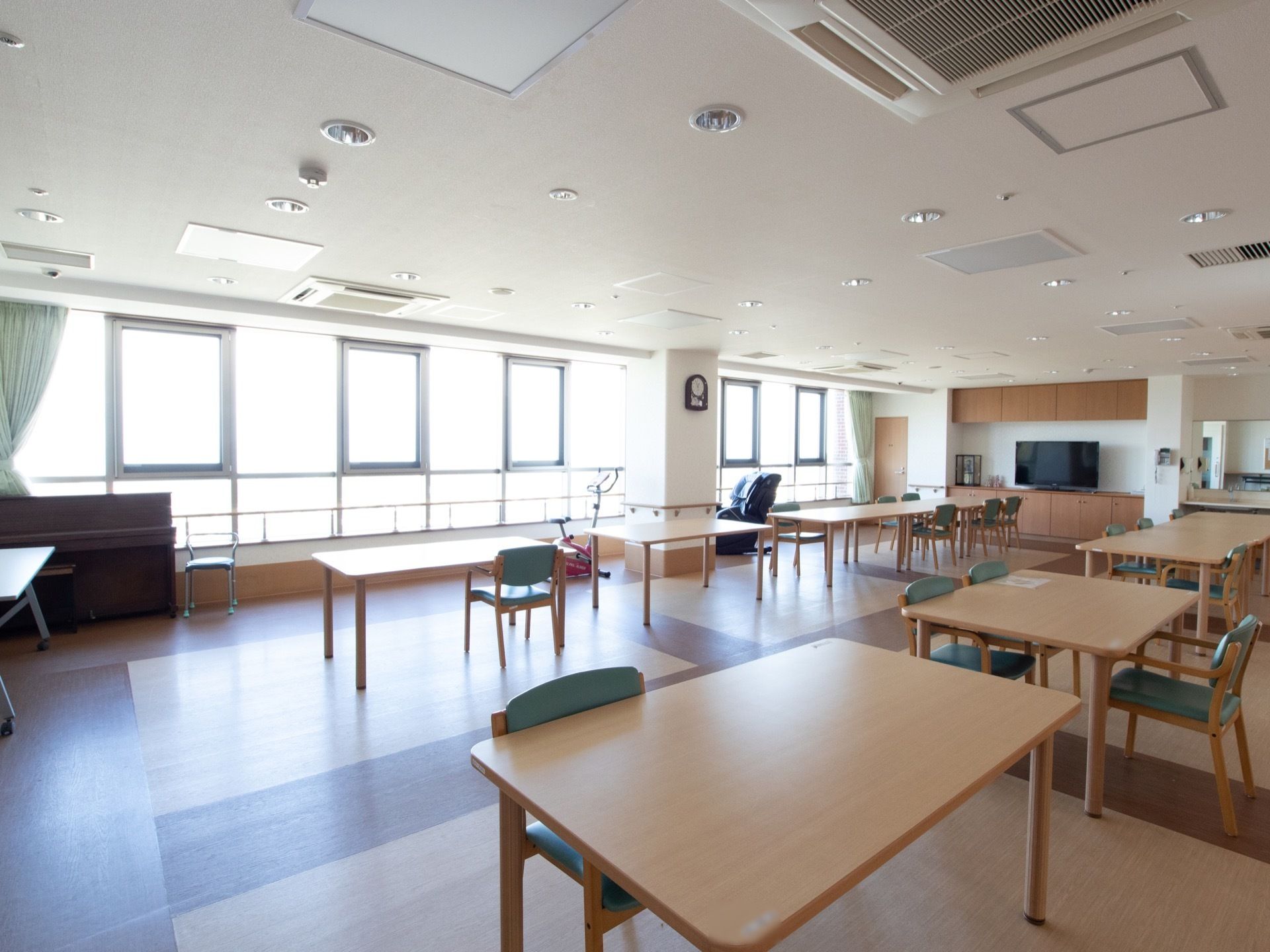 部屋の１面が窓ガラスになっていて、室内がとても明るい。たくさんのダイニングテーブルと椅子があり、壁に時計が掛けられている。