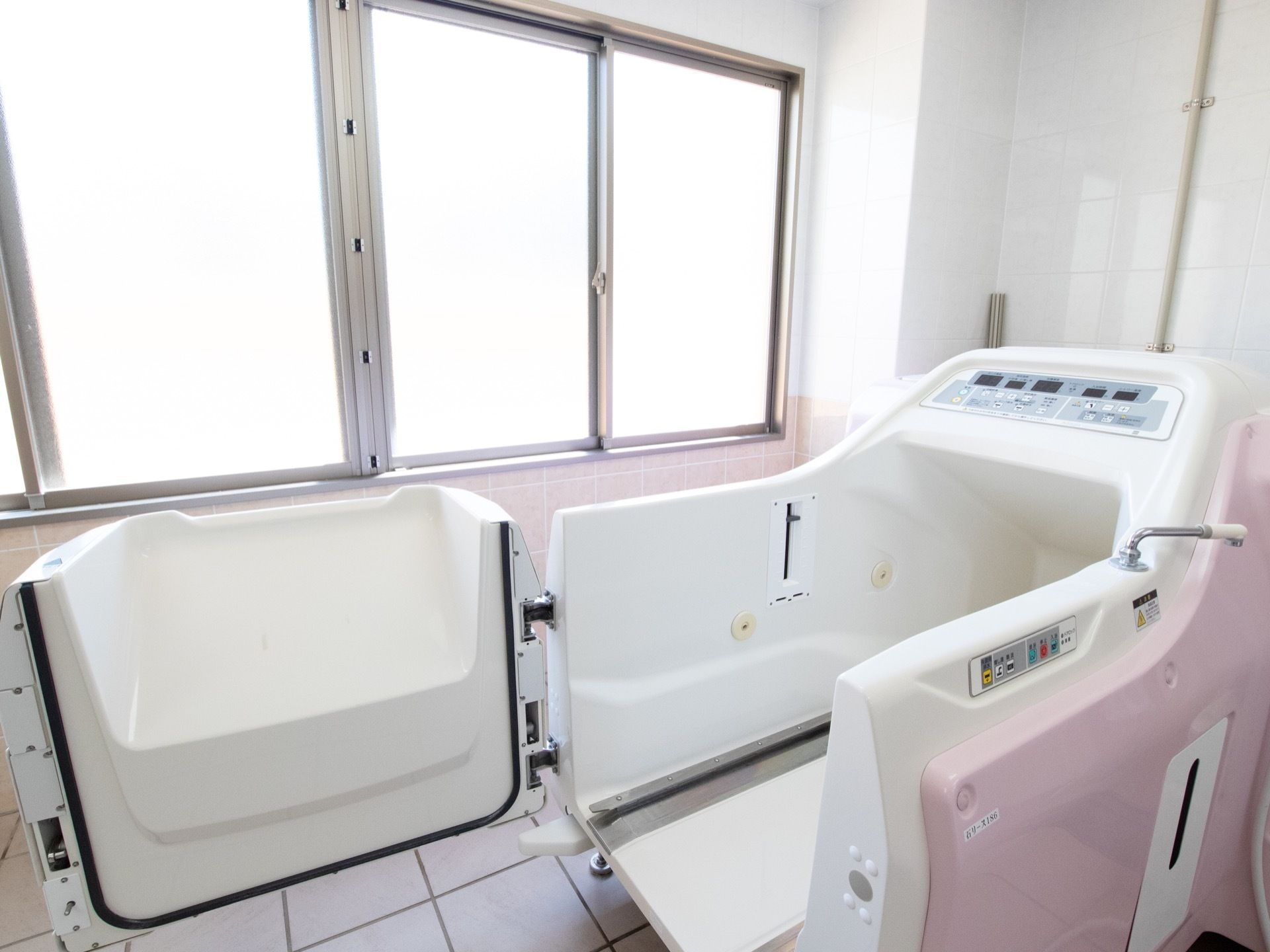 壁に白色と薄いピンク色のタイルと床は白いタイルが貼られた浴室である。白色とピンク色の介護浴槽の扉が開かれている。