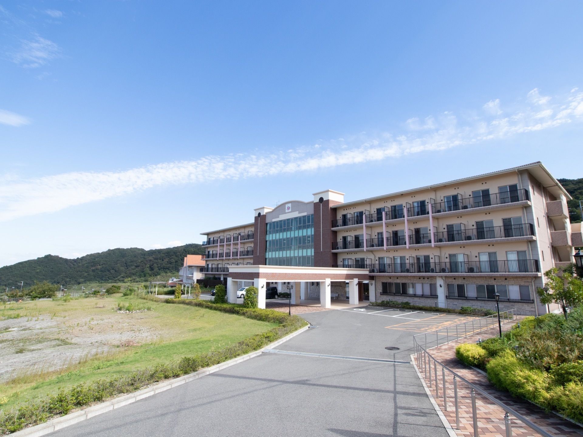 学校の校舎のような４階建ての建物の奥に山があり、青空には雲がかかっている。敷地の前には広々とした緑の土地がある。