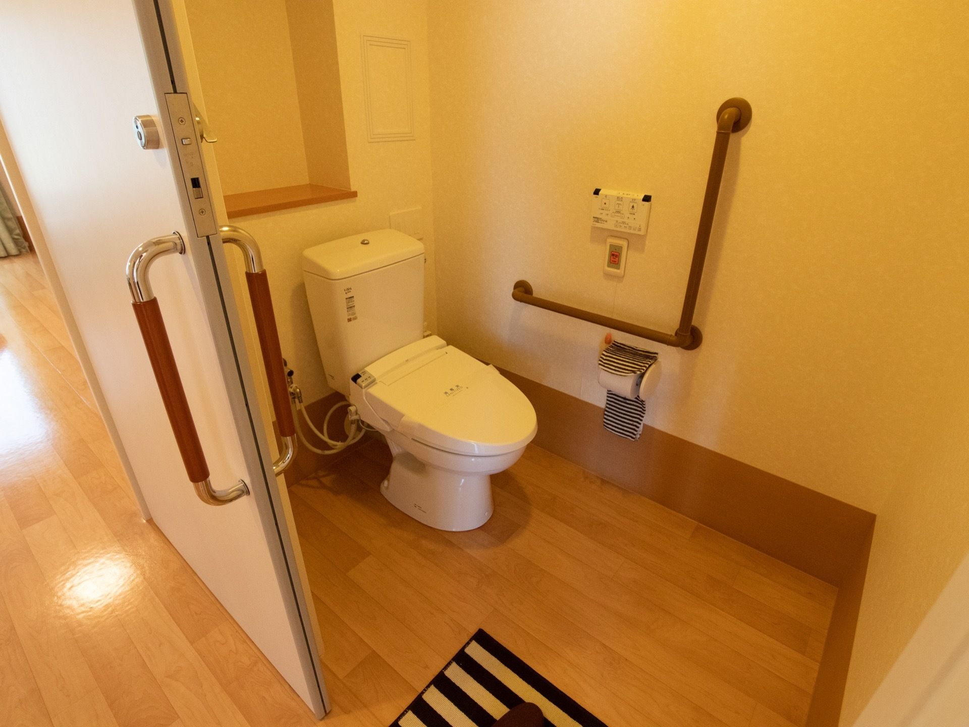 フローリングの床にトイレマットが敷いてある。壁に手すりとナースコールが設置され、ペーパーホルダーにストライプのカバーが掛けられている。