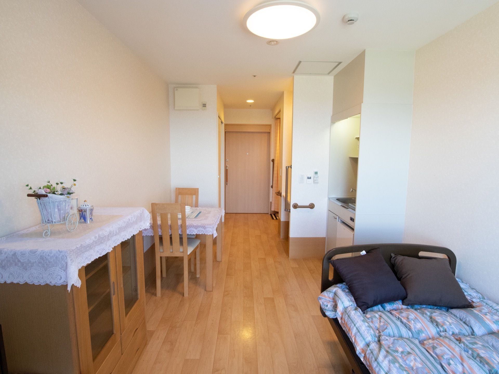 縦長の部屋にベッドとダイニングセットと収納棚が置いてある。ベッドの上にはクッションが２つあり、天井にシーリングライトが設置されている。