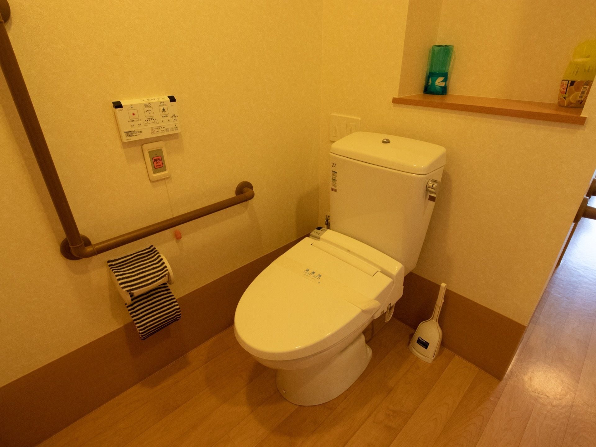 タンク付きの白い洋式便器の下にトイレブラシが置いてある。棚の上に２種類の消臭剤があり、壁に温水洗浄便座のリモコンが設置されている。