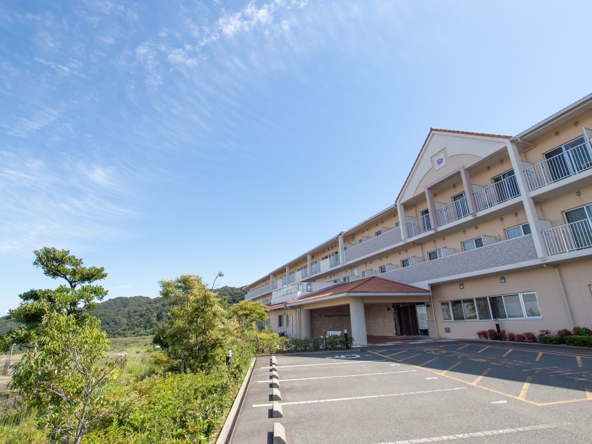 澄み渡った青空に薄いすじ雲が掛かっている。３階建ての建物の前にアスファルトで整備された駐車スペースがある。