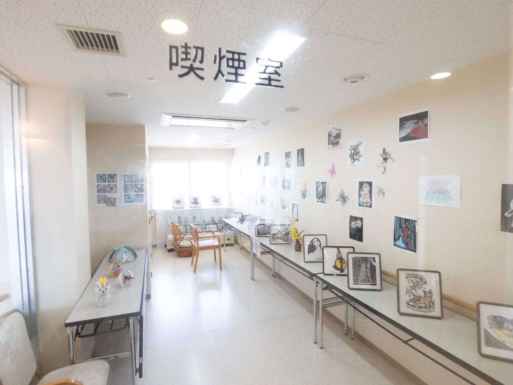 透明の板に喫煙室と黒文字で表示されている。細長い部屋の両側に長テーブルが置いてあり、壁に絵などが飾られている。