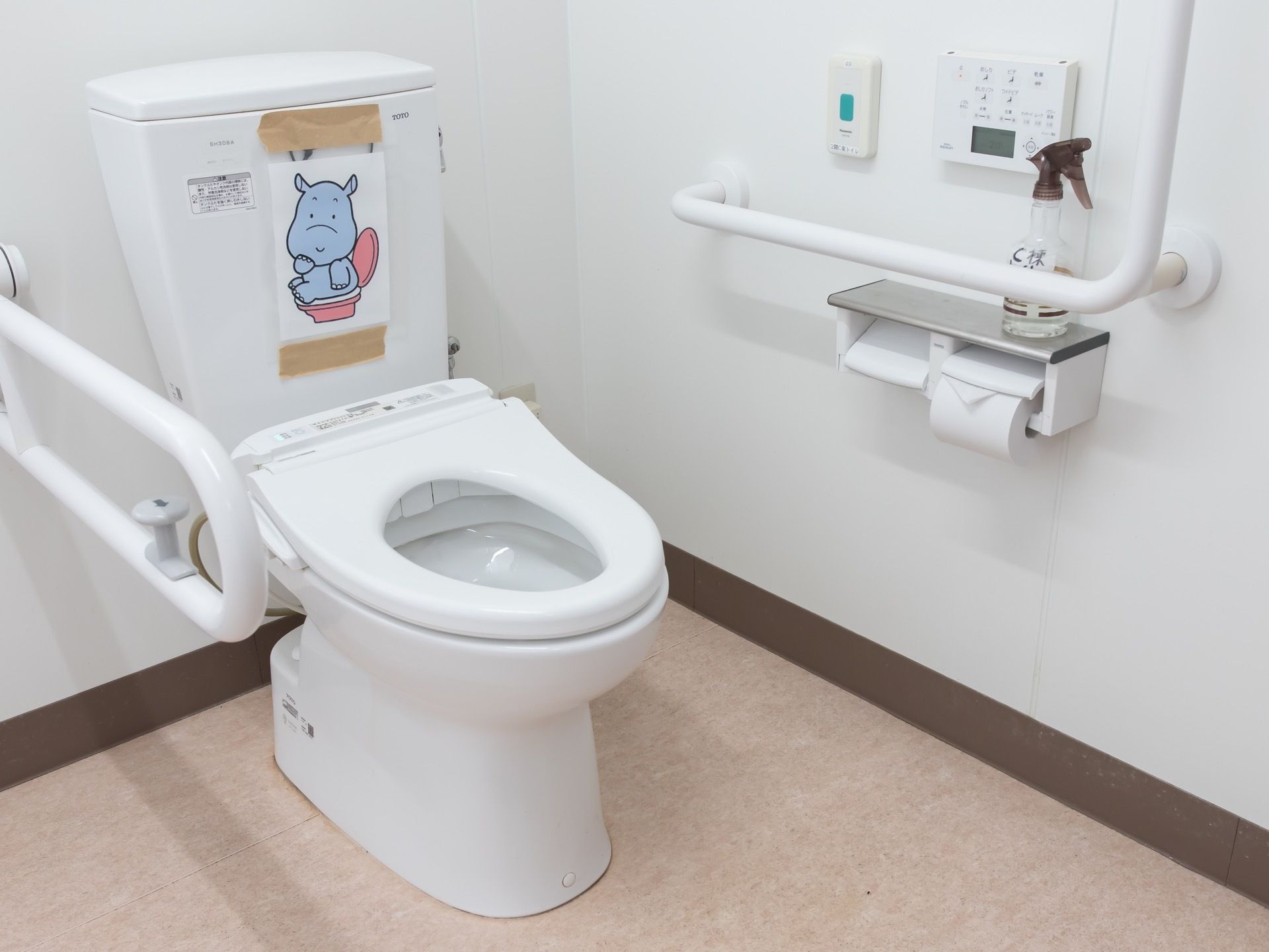 白い壁で、床にベージュのシートが張られたトイレに、洗浄機能付きの便座が据えられている。右の壁面と便座の左側に手すりが付けられている。