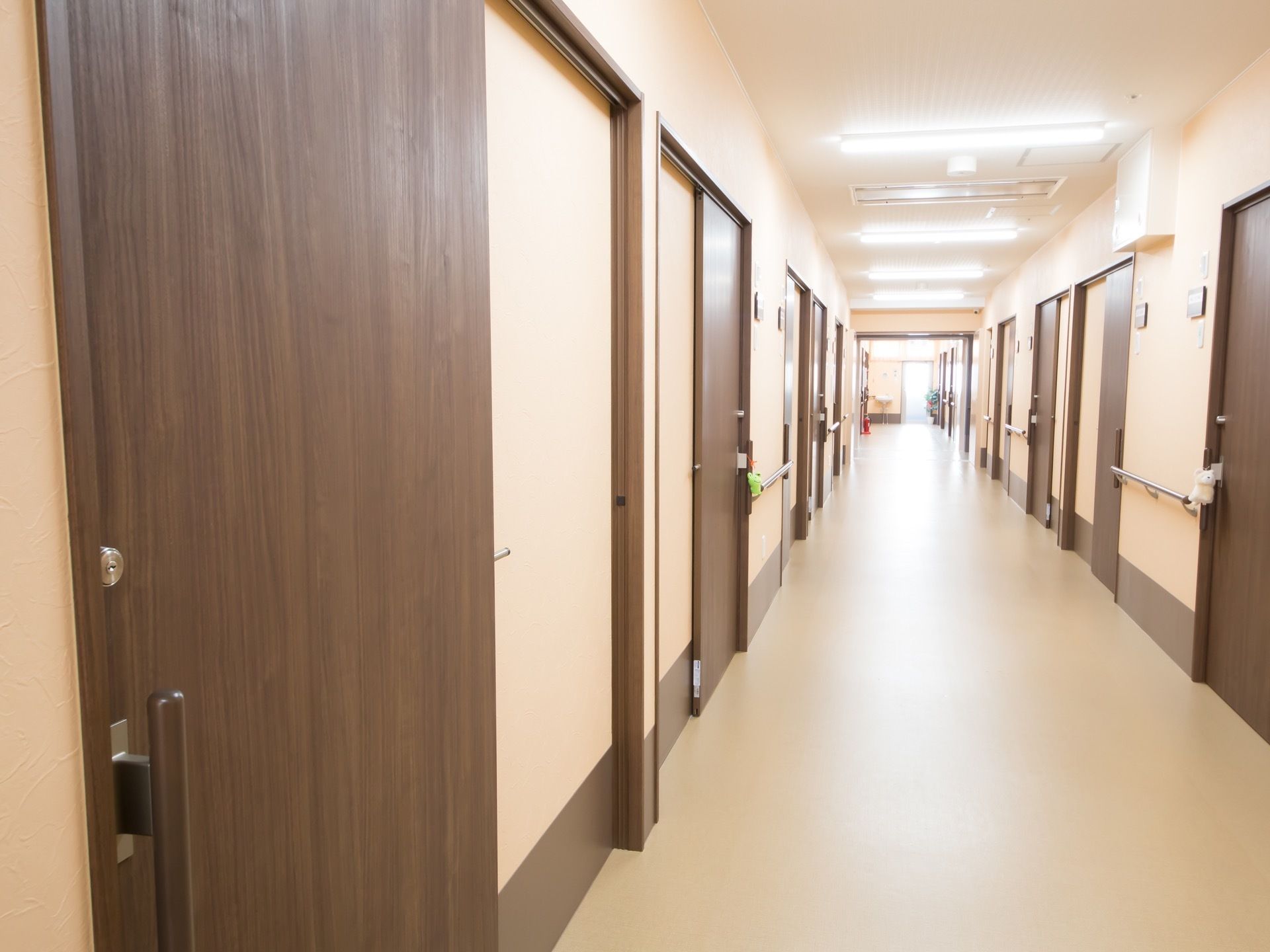 ダークブラウンの居室のドアが並ぶ廊下がある。壁と床はあたたかみのある色調となっている。壁には手すりと表札がついている。