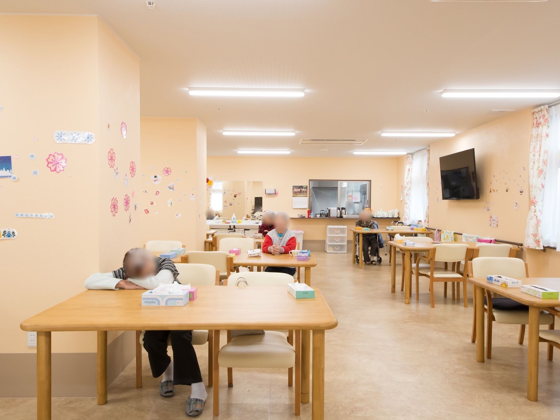 ベージュの壁の明るい食堂がある。テーブルと椅子が並べられ、大きな柱が立っている。壁にはカーテンの掛かった窓とテレビが設置されている。