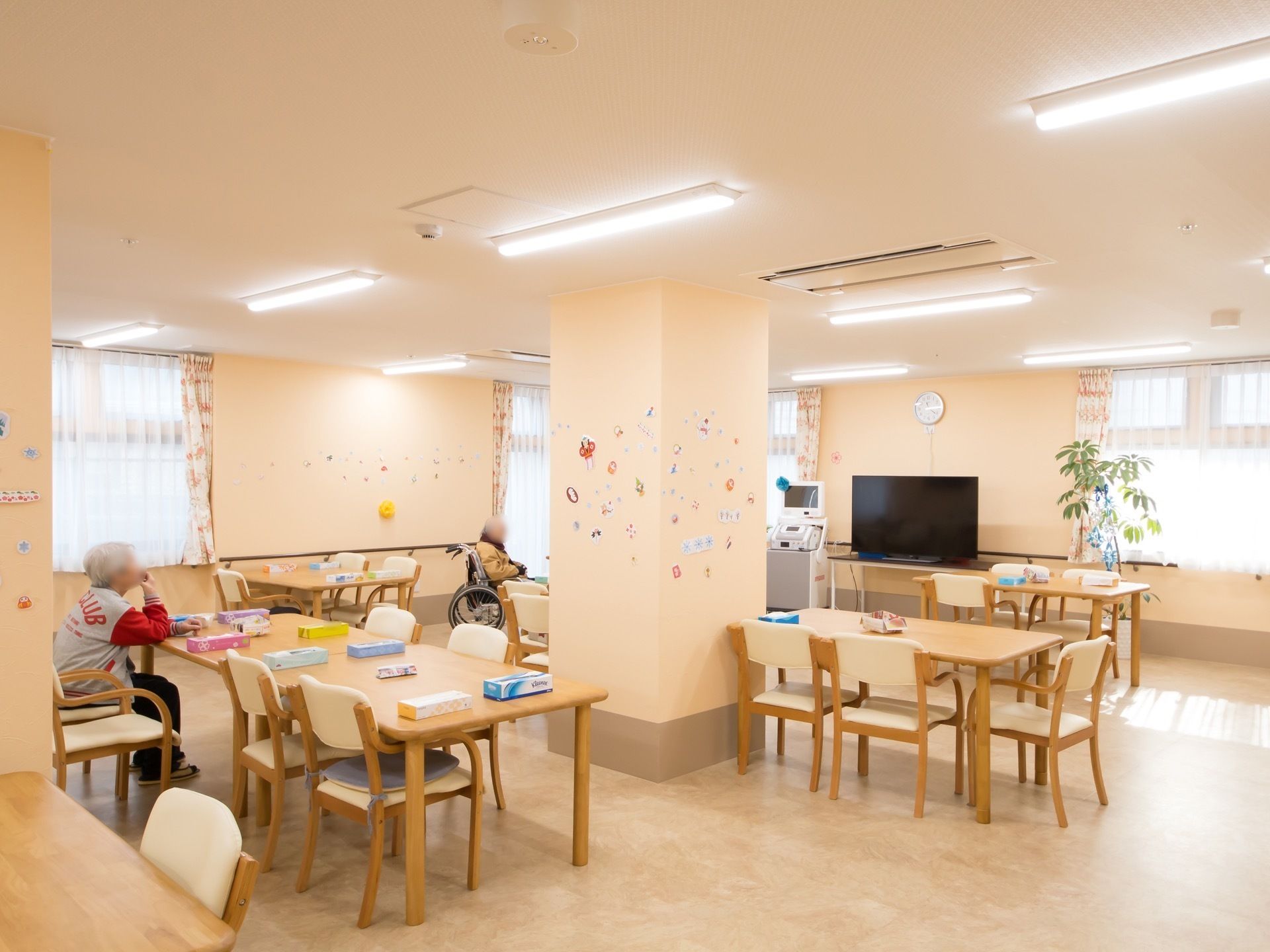 クリーム色の壁の明るく広い食堂がある。カーテンの掛かった窓があり、テーブルが並べられている。壁には手すりと大きなテレビが置かれている。