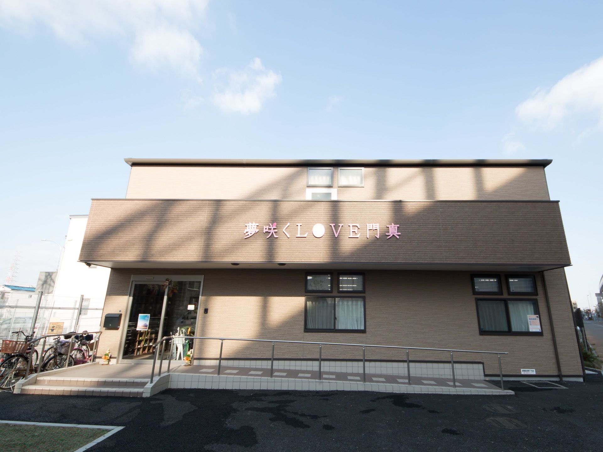 ベージュのタイルの壁に囲まれた２階建ての建物がある。玄関先には２段の階段と、手すりのついたスロープが設置されている。