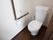 ダークブラウンの床と白い壁のトイレがある。壁にはL字型の手すりが設置され、緊急用ブザーがついている。