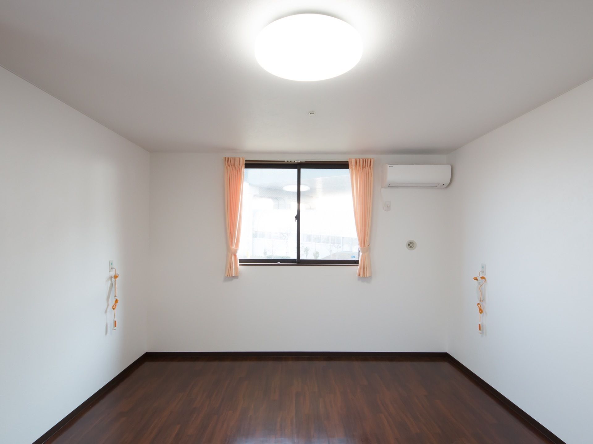 木目調のダークブラウンの床と白い壁の明るい居室がある。大きな窓があり、カーテンが掛かっている。エアコンが設置されている。