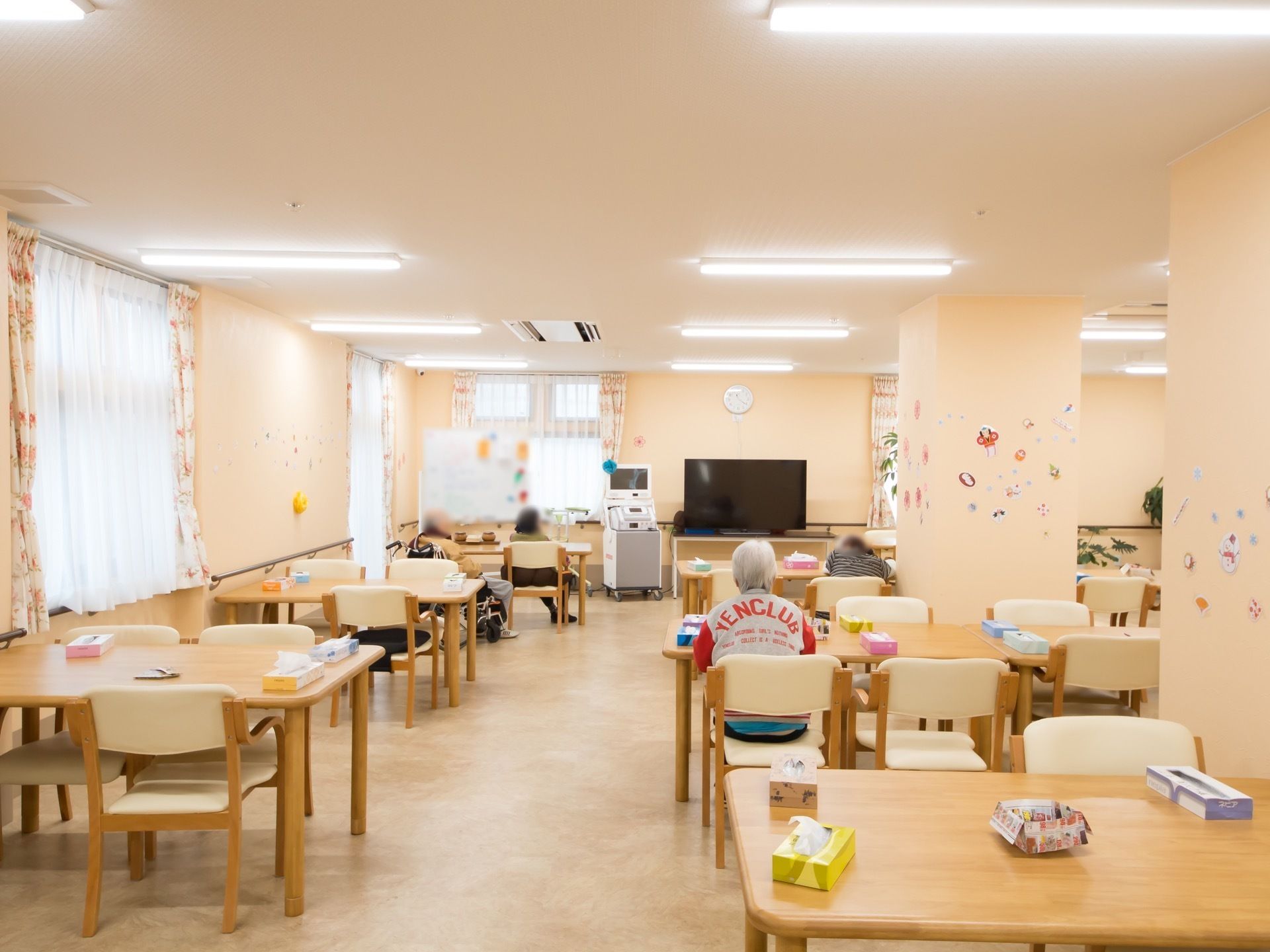 テーブルと椅子がたくさん並んでいる広い食堂がある。大きなテレビが置かれている。壁には手作りの飾りつけがされている。