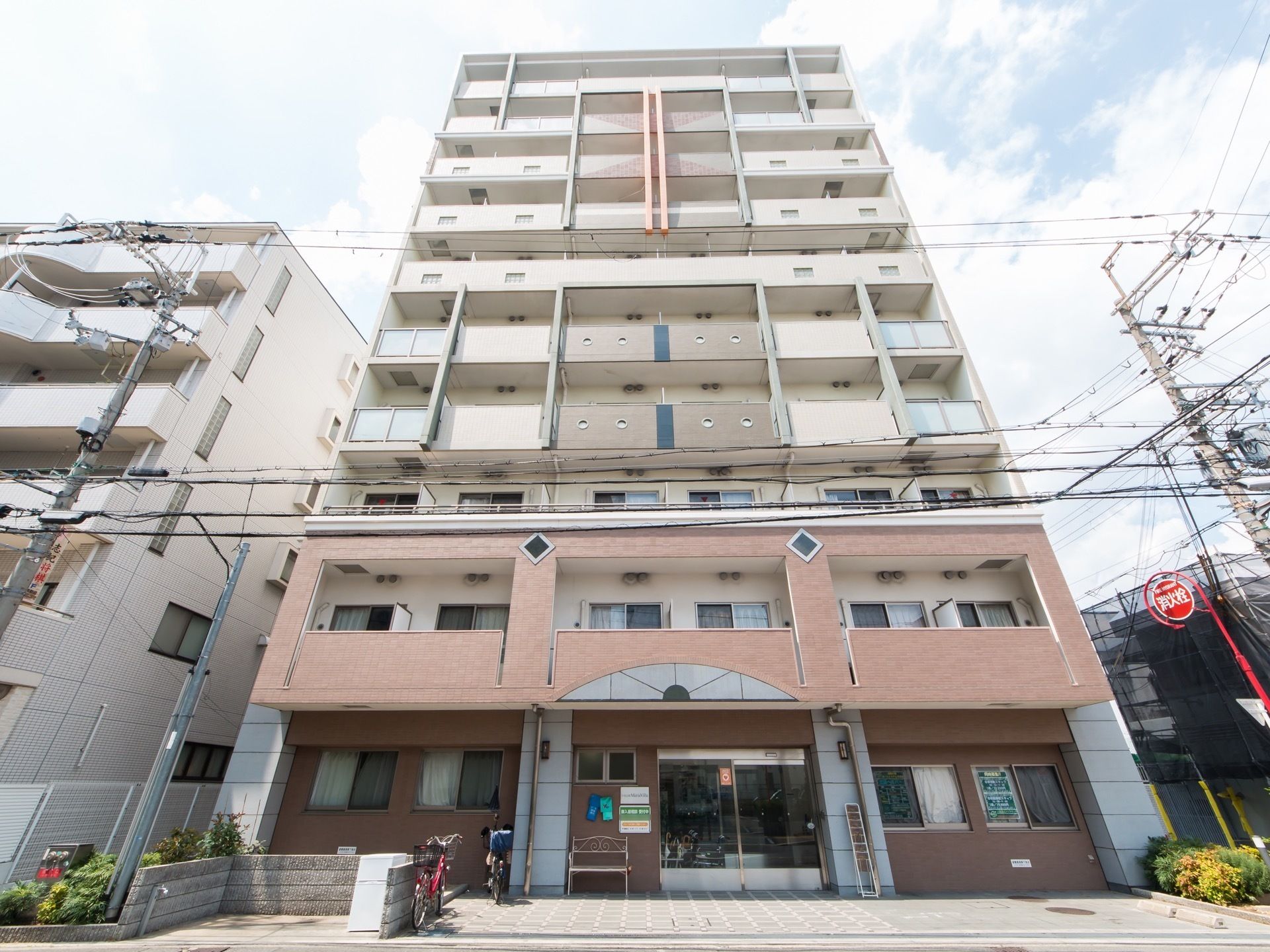 施設は高い建物で、玄関の横にはベンチや自転車が置かれている。2階以上にはベランダが付いており、白い仕切り板が見える。