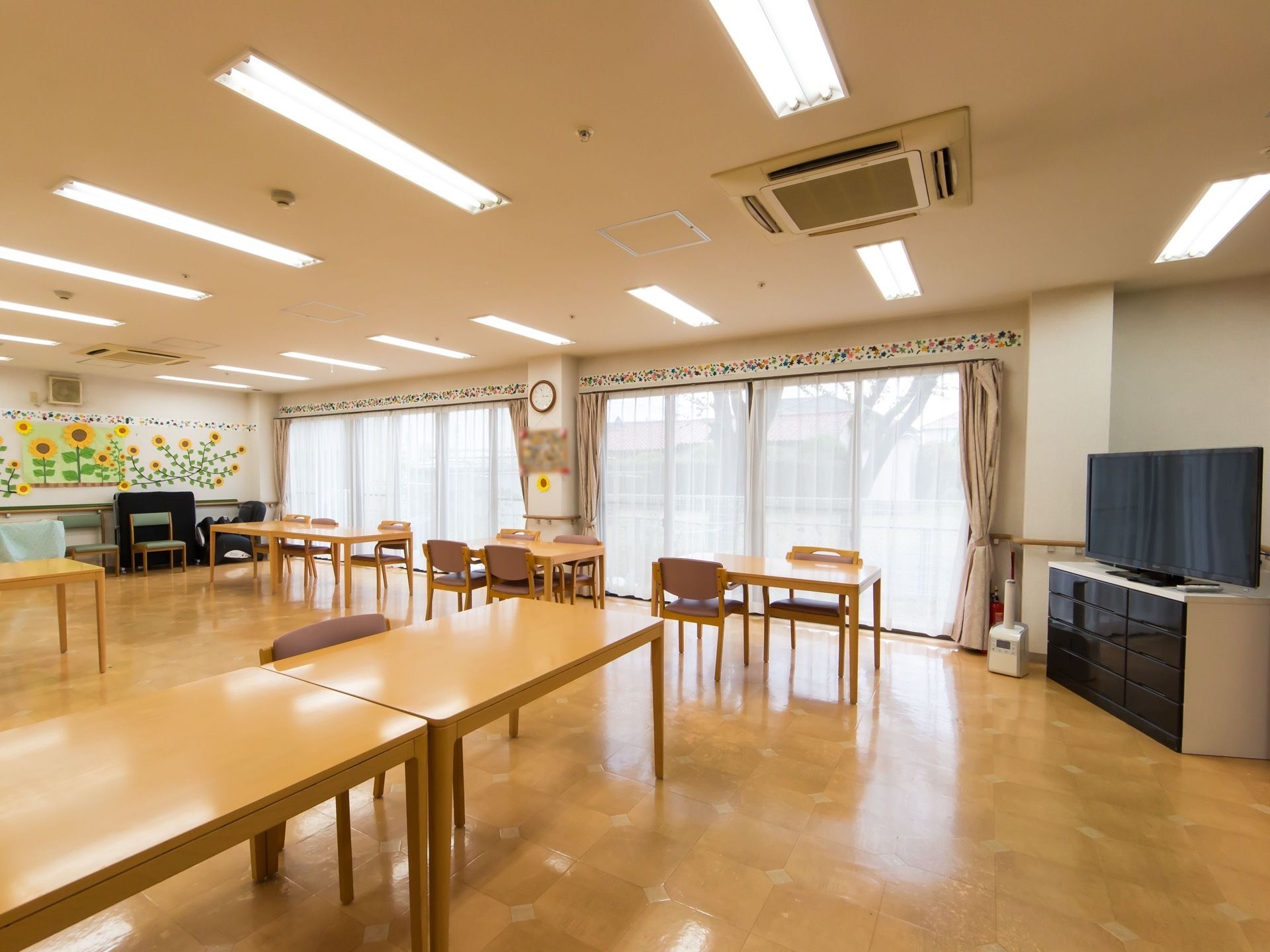 掃き出し窓のある明るい食堂。天井には空調が付いていて、フロアにはテーブルセットやテレビが置かれている。