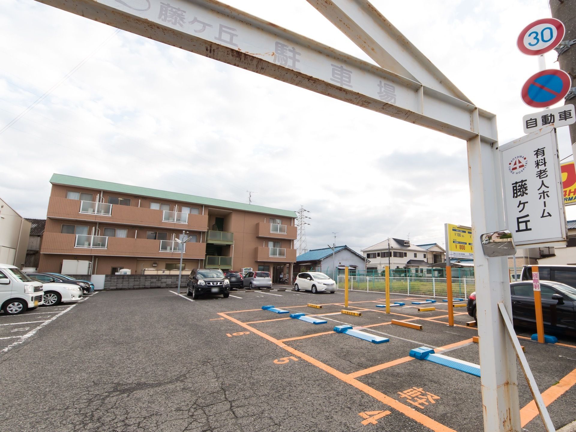 アスファルトで舗装された駐車場があり、一部はコインパーキングになっている。奥には、茶色い外壁の３階建て住宅が建っている。