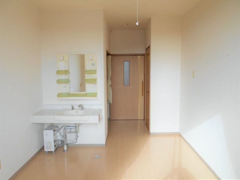 車いす対応の洗面所が付いたシンプルな部屋。出入り口付近にはカーテンで開け閉めするタイプのトイレがある。