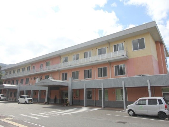 広い駐車場が付いた3階建ての建物。ベージュとサーモンピンクの外壁で明るい雰囲気。