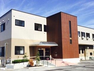 ブラウン系の２階建ての建物。玄関には手すり付きのスロープと屋根が付いている、草木も植えられている。