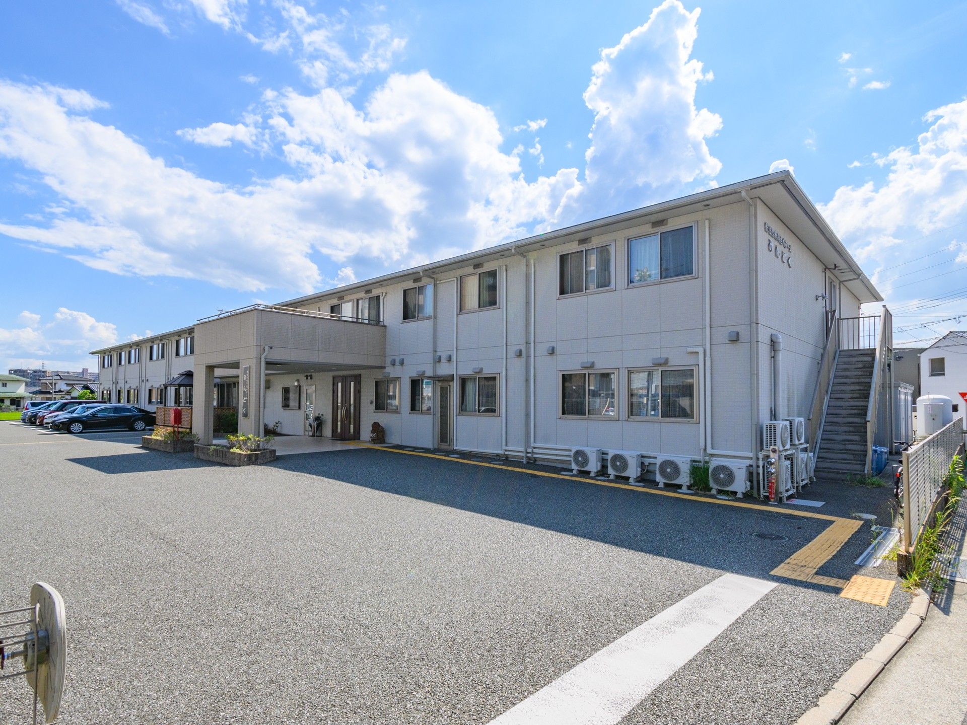 玄関周辺には木が植えられ、看板も掲げられている。温かみのあるベージュの建物。