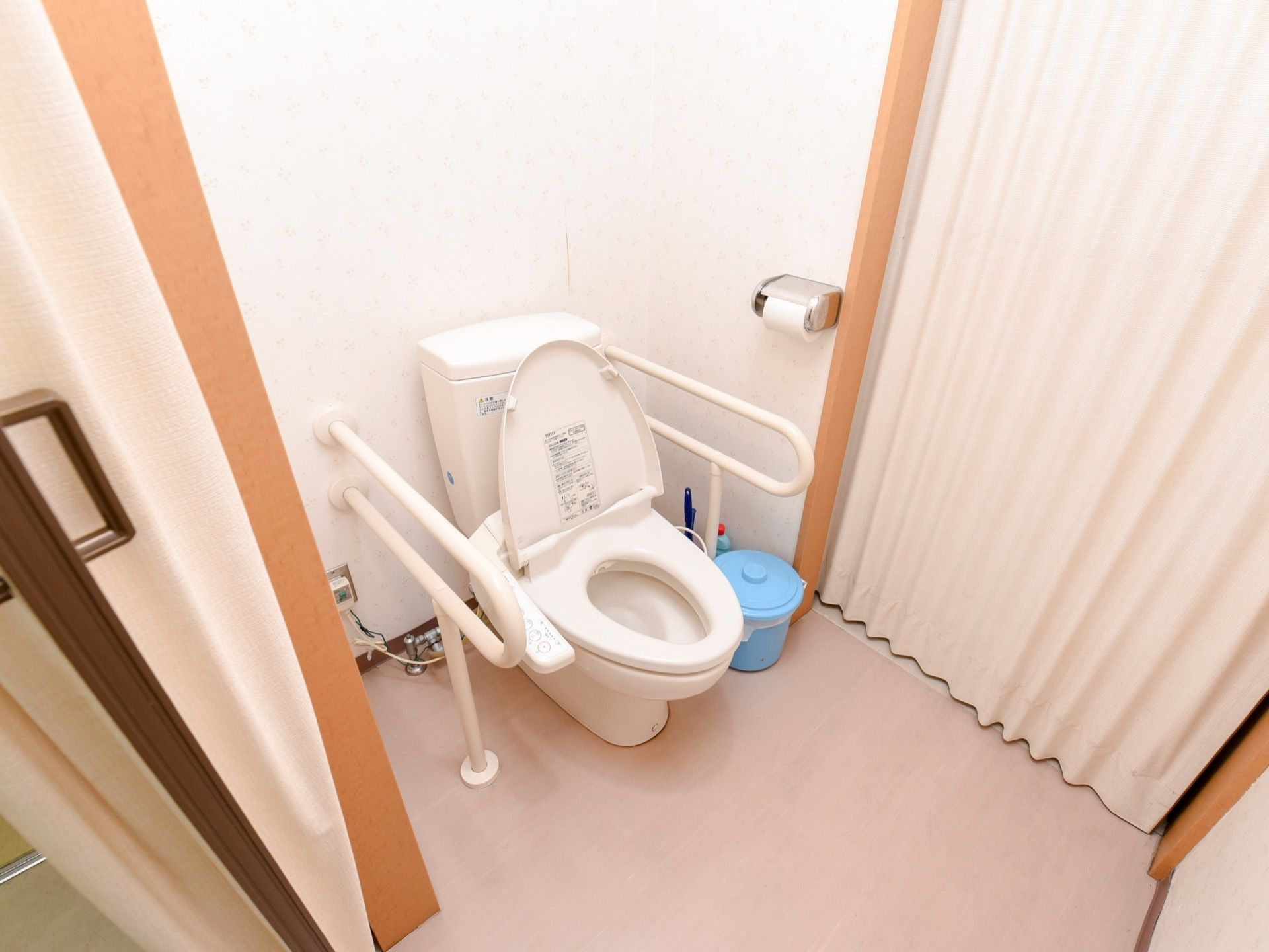 トイレはアコーディオンカーテンで区切られている。便器の両側に白い手すりが設置されている。便座は洗浄機能付きである。