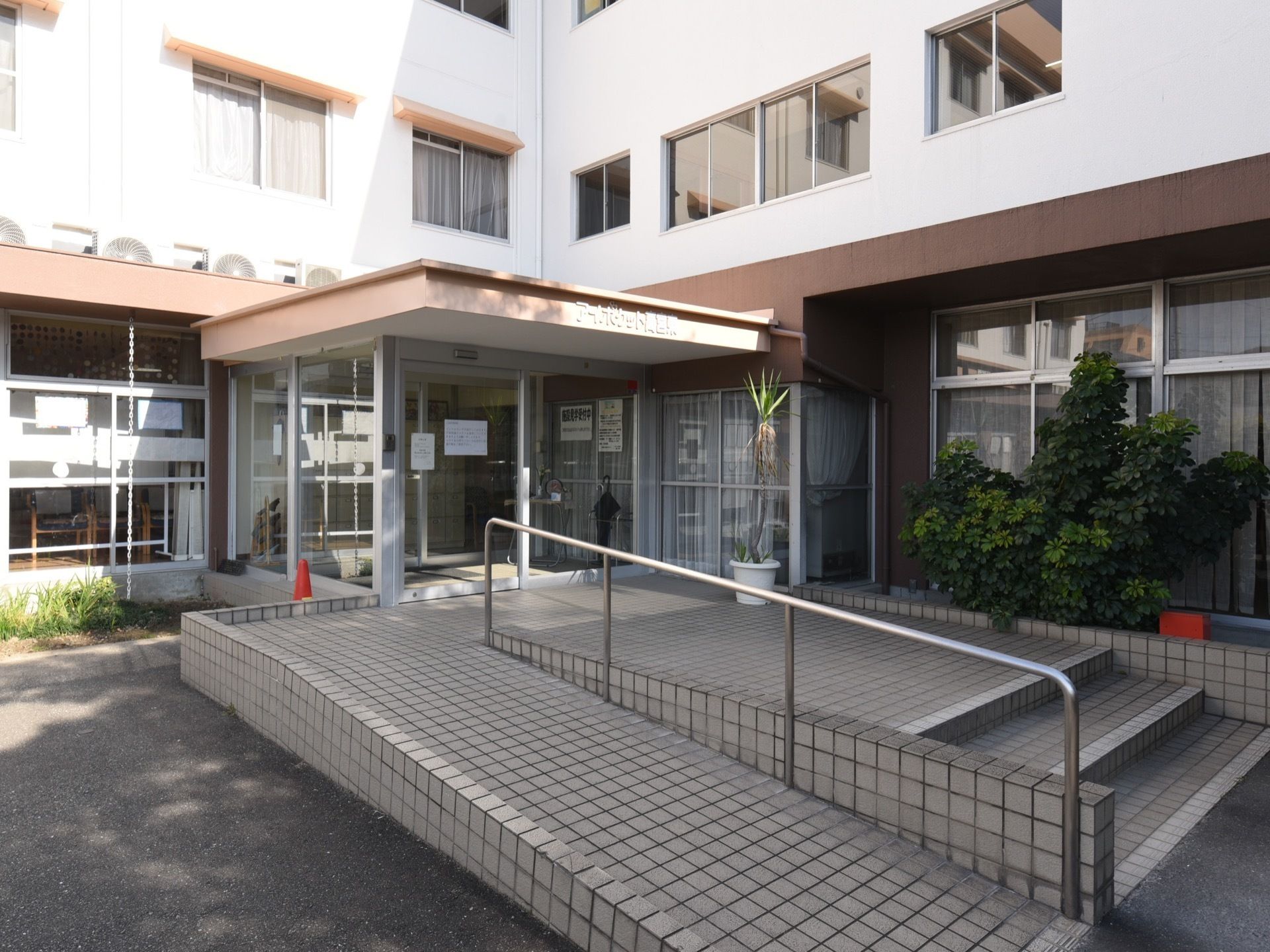 ２面の壁に囲まれているガラス張りの入口がある。２枚のドアの前には、階段とスロープが並んで設置してある。
