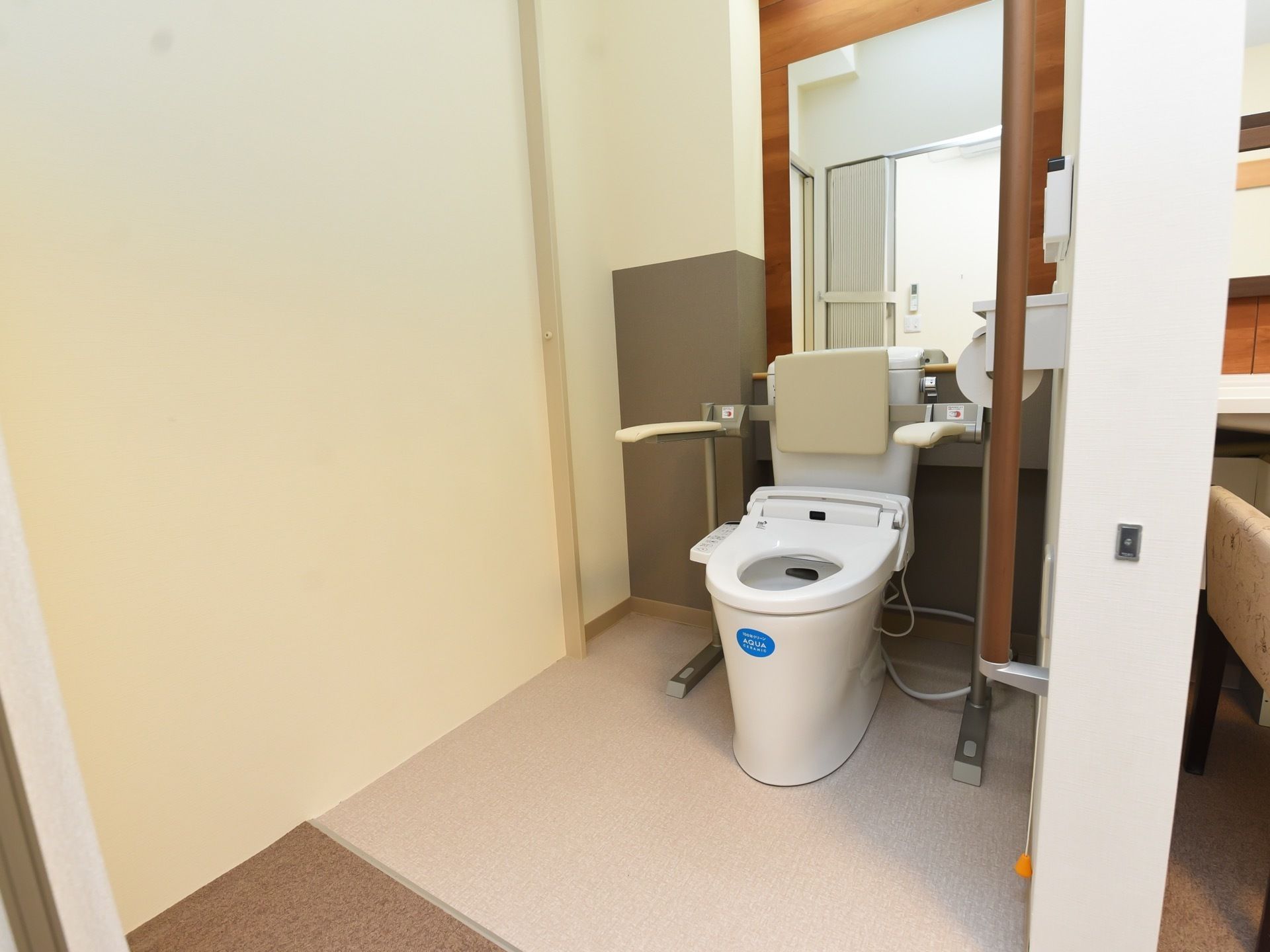 白色の洋式便座を設置している。背後に縦型の鏡が備えつけられている。両サイドに手すり、背もたれもある。