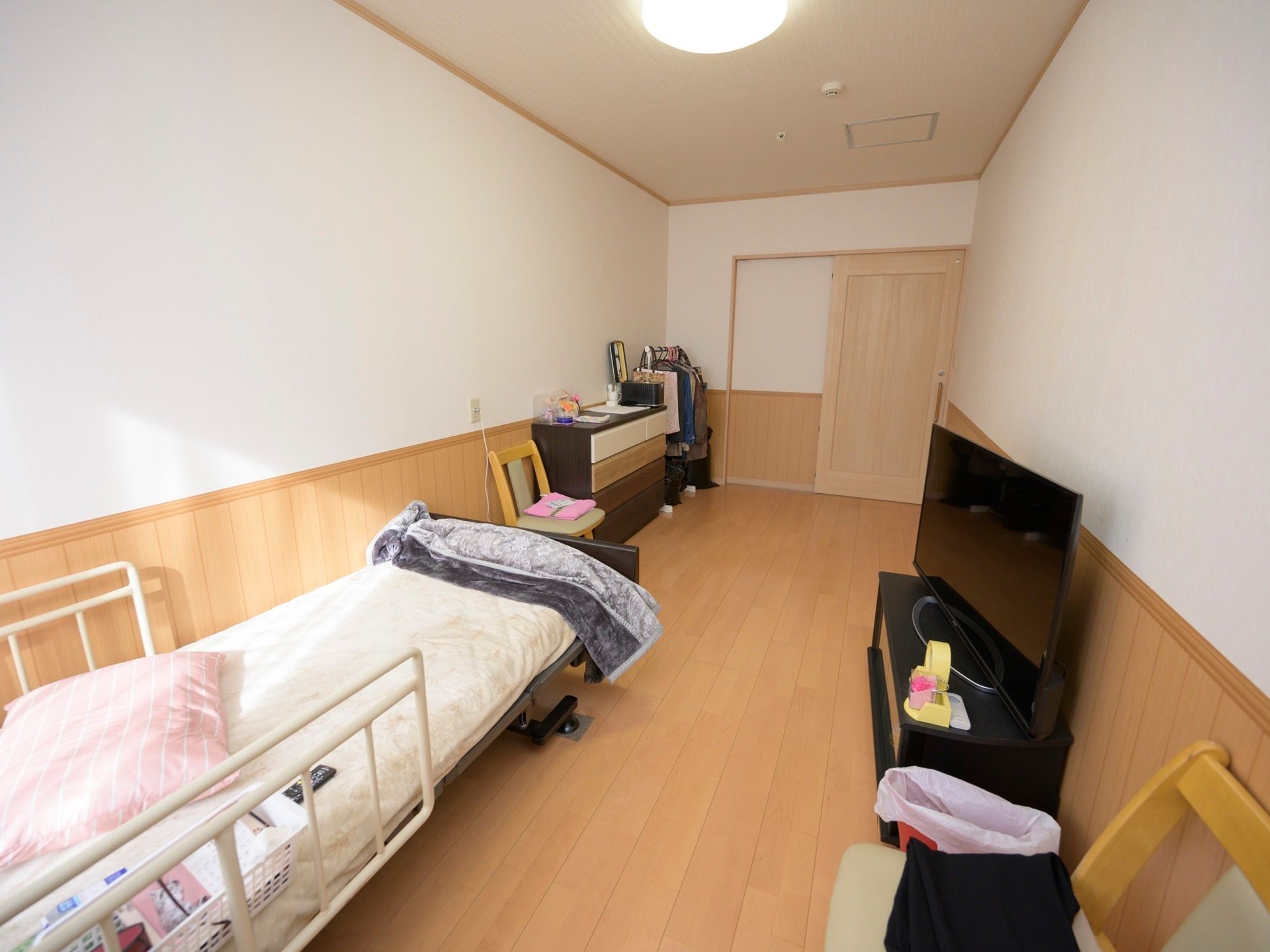 白い壁に明るい木目調を基調に整えられている居室である。テレビや家具類、介護用ベッドが設置されている。