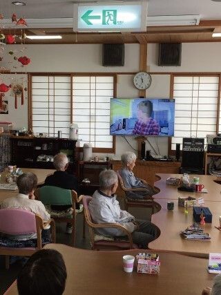 大きなテレビに映っている画像を熱心に見ている高齢者たち。複数のダイニングテーブルに分かれて座っている。