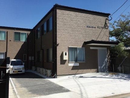 エル字型の2階建ての建物はブラウン系で駐車場には車が止まっていて、玄関には屋根も付いている。