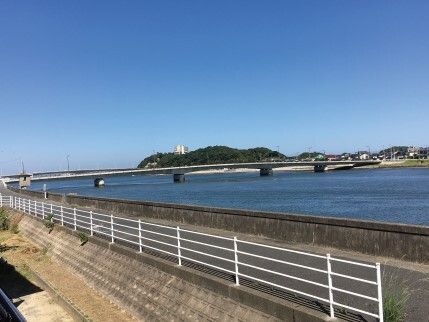 川に面した道路。川には多くの水が流れていて、橋もかかっている。橋の向こうには山や住宅地もある。
