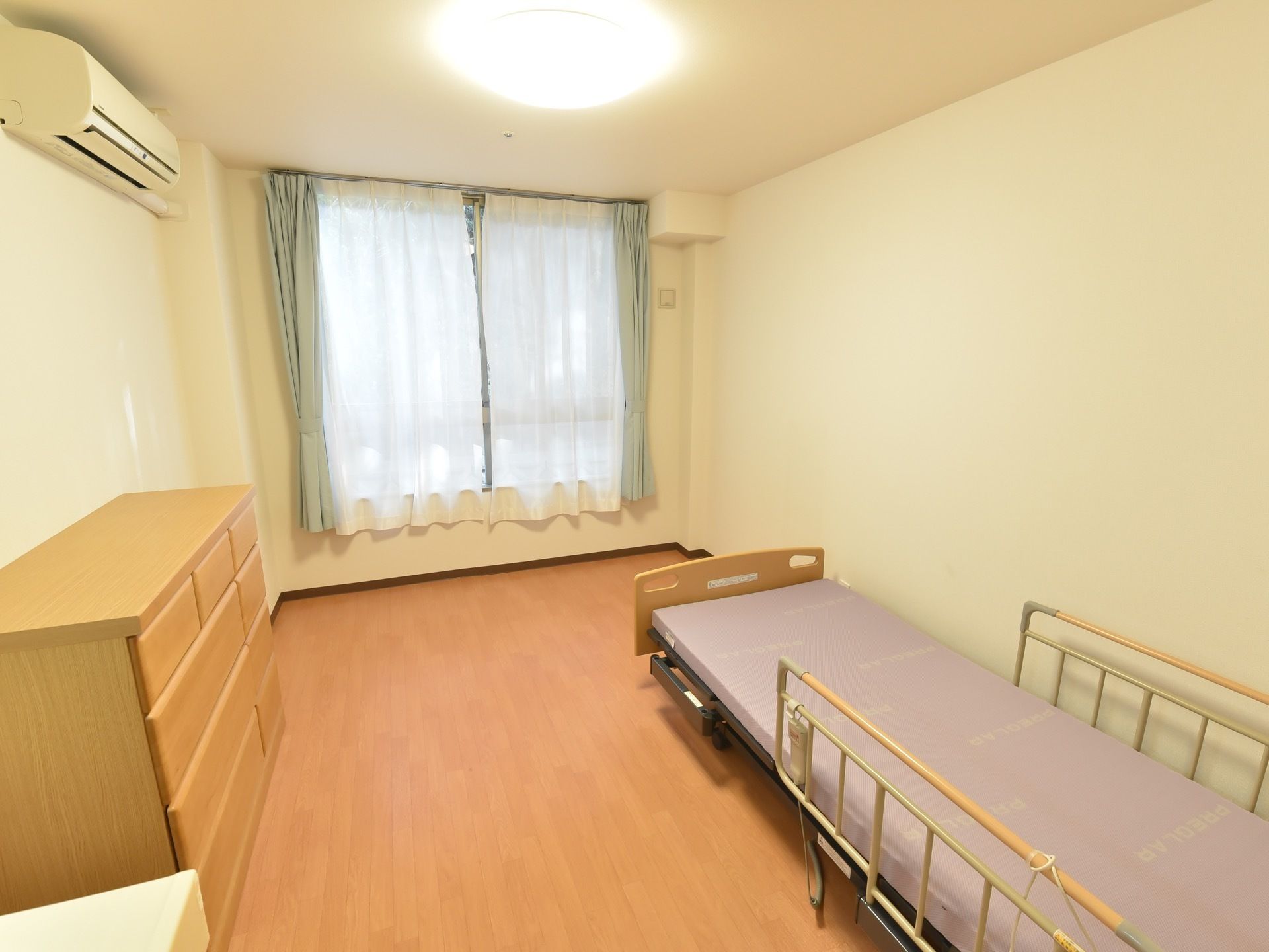 窓やエアコンが完備されている居室である。木目調のフローリングに、整理ダンスや介護用ベッドが設置されている。