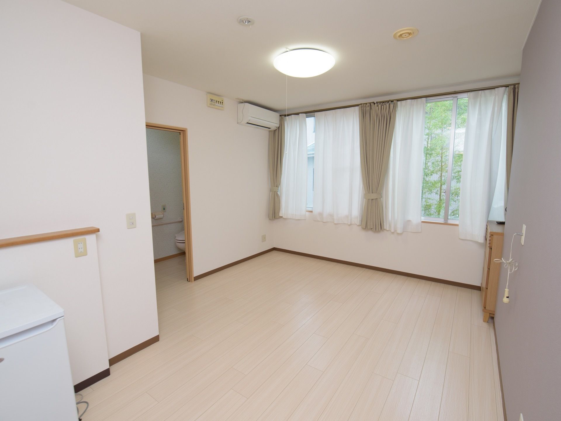 エアコンが完備された居室である。窓にはカーテンが掛けられている。壁の中央にあるコンセントにはナースコールが取り付けられている。