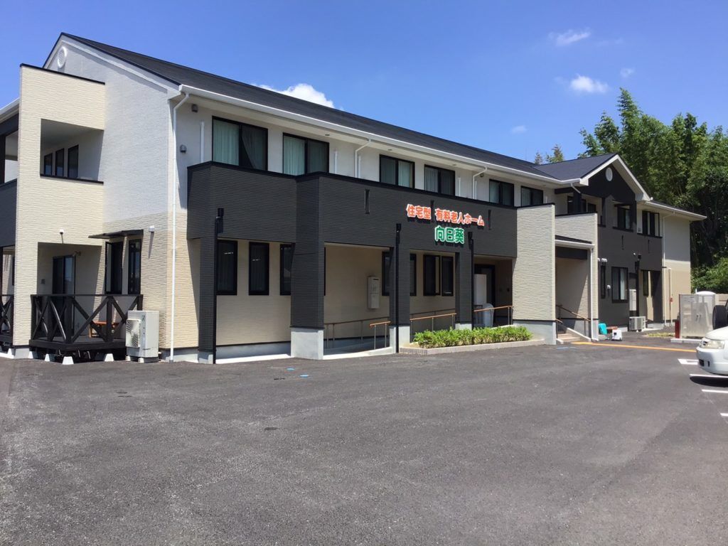 手すり付きのスロープと階段がある玄関。2階建ての建物は黒とベージュで、ウッドデッキのテラスもある。