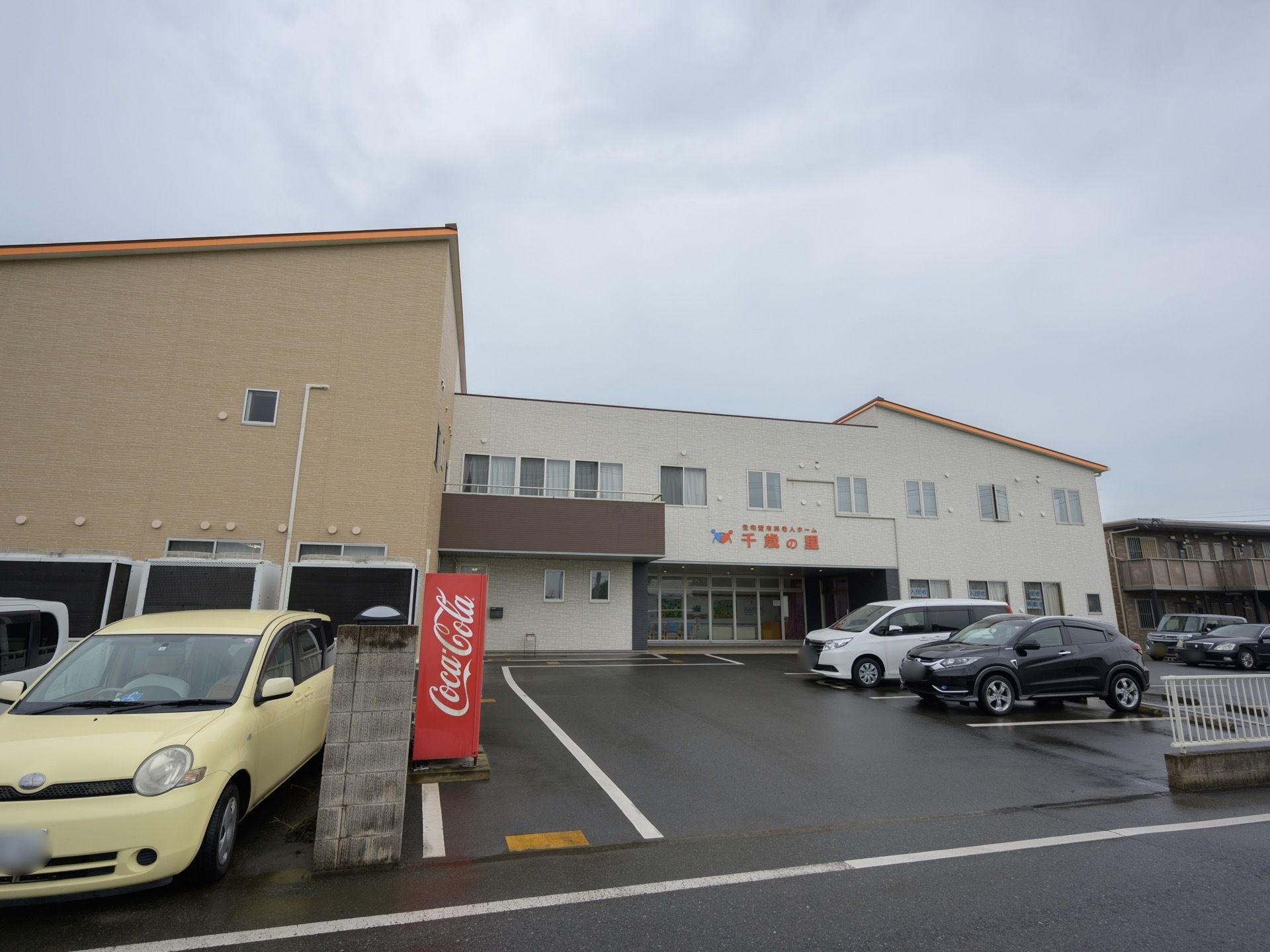 舗装され、白線で区分された駐車場が、２階建ての施設の前にある。建物の中央部にある玄関上に赤字で施設名が記載されている。
