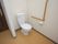 部屋の入り口近くに設けられたトイレである。ドアはない。温水洗浄便座が設置され、壁にはＬ字型の手すりとペーパーホルダーが取り付けてある。