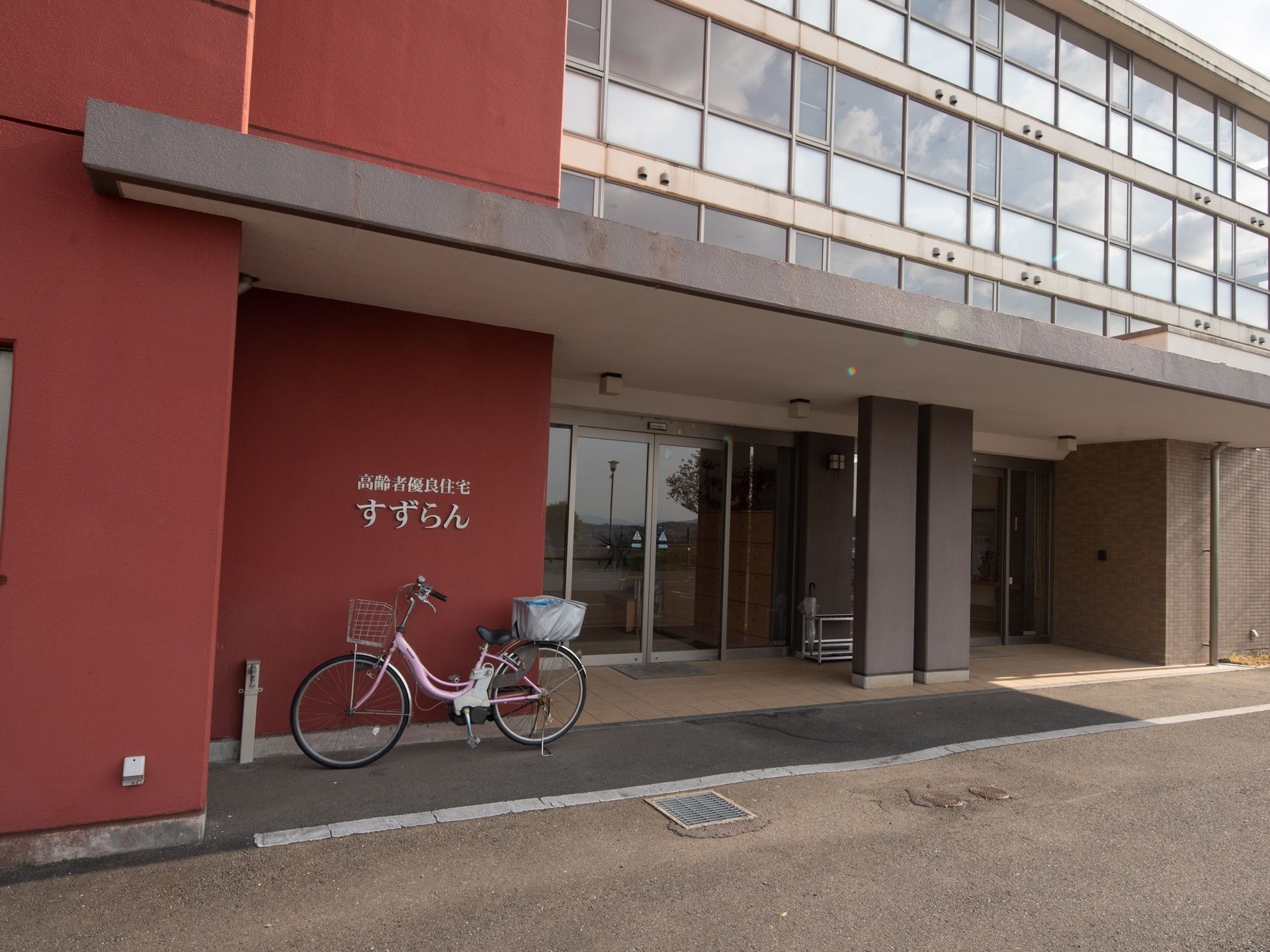 施設入口に自動扉が完備されている。横のレンガ色の外壁には、施設名称と施設種別が提示され、前には1台の自転車が止められている。